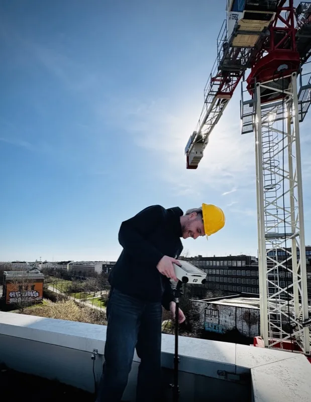 Teammitglied mit Schutzhelm richtet Stativ auf dem Dach aus